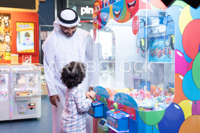مشاركة الأب ابنه لحظات المرح والحماس ، تعابير السرور والسعادة، طفل خليجي يرتدي الزي الكاجوال يقوم  بلعب على اله المخلب، قضاء أوقات ممتعة باللعب والتركيز، أجواء تفاعلية مبهجة ، رجل عربي خليجي اماراتي يرتدي الكندورة و الغتره يستمتع بقضاء وقت ممتع مع طفله فى مدينه الملاهي