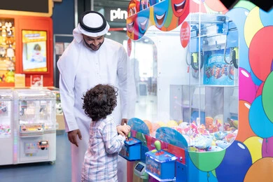 مشاركة الأب ابنه لحظات المرح والحماس ، تعابير السرور والسعادة، طفل خليجي يرتدي الزي الكاجوال يقوم  بلعب على اله المخلب، قضاء أوقات ممتعة باللعب والتركيز، أجواء تفاعلية مبهجة ، رجل عربي خليجي اماراتي يرتدي الكندورة و الغتره يستمتع بقضاء وقت ممتع مع طفله فى مدينه الملاهي