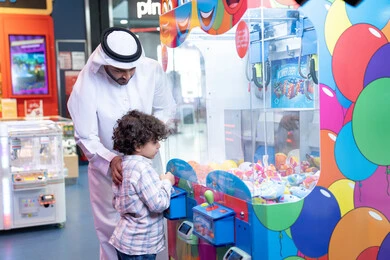 تعابير السرور والسعادة، طفل خليجي يرتدي الزي الكاجوال يقوم  بلعب على اله المخلب، قضاء أوقات ممتعة باللعب والتركيز، مشاركة الأب ابنه لحظات المرح والحماس ، أجواء تفاعلية مبهجة ، رجل عربي خليجي اماراتي يرتدي الكندورة و الغتره يستمتع بقضاء وقت ممتع مع طفله فى مدينه الملاهي