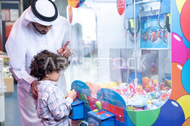 طفل خليجي يرتدي الزي الكاجوال يقوم  بلعب على اله المخلب، قضاء أوقات ممتعة باللعب والتركيز،  تعابير السرور والسعادة، مشاركة الأب ابنه لحظات المرح والحماس ، أجواء تفاعلية مبهجة ، رجل عربي خليجي اماراتي يرتدي الكندورة و الغتره يستمتع بقضاء وقت ممتع مع طفله فى مدينه الملاهي