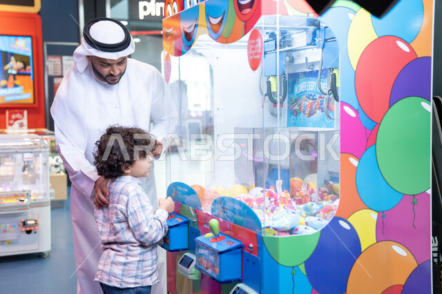 A fun atmosphere full of fun and imagination, a Gulf child wearing casual clothes playing on the claw machine, expressions of pleasure and happiness, the father sharing moments of fun and enthusiasm with his son, a cheerful interactive atmosphere, an Arab Gulf Emirati man wearing a kandura and ghutra enjoying a fun time with his child in the amusement park