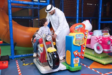 الاستمتاع بتجربة لعب مميزة وآمنة، رجل عربي خليجي اماراتي يرتدي الكندورة و الغتره يستمتع بقضاء وقت ممتع مع ابنه فى مدينه الملاهي،  طفل عربي خليجي يركب لعبة الدراجه المتحركه بايماءات السعاده و السرور، التفاعل بين أفراد العائلة، الأنشطة  الترفيهيه الممتعة