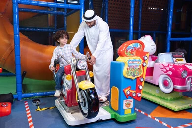 مركز العاب ترفيهي داخلي، رجل عربي خليجي اماراتي يرتدي الكندورة و الغتره يستمتع بقضاء وقت ممتع مع ابنه فى مدينه الملاهي،  طفل عربي خليجي يركب لعبة الدراجه المتحركه بايماءات السعاده و السرور، التفاعل بين أفراد العائلة، الأنشطة  الترفيهيه الممتعة