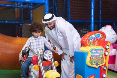 أجواء مليئة بالمرح والحماس، رجل عربي خليجي اماراتي يرتدي الكندورة و الغتره يستمتع بقضاء وقت ممتع مع ابنه فى مدينه الملاهي،  طفل عربي خليجي يركب لعبة الدراجه المتحركه بايماءات السعاده و السرور، التفاعل بين أفراد العائلة، الأنشطة  الترفيهيه الممتعة