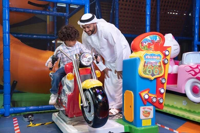 رجل عربي خليجي اماراتي يرتدي الكندورة و الغتره يستمتع بقضاء وقت ممتع مع ابنه فى مدينه الملاهي،  طفل عربي خليجي يركب لعبة الدراجه المتحركه بايماءات السعاده و السرور، التفاعل بين أفراد العائلة، الأنشطة الرياضية الممتعة، أجواء المرح والحماس