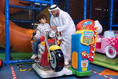 طفل عربي خليجي يركب لعبة الدراجه المتحركه بايماءات السعاده و السرور، التفاعل بين أفراد العائلة، الأنشطة الرياضية الممتعة، أجواء المرح والحماس ، رجل عربي خليجي اماراتي يرتدي الكندورة و الغتره يستمتع بقضاء وقت ممتع مع ابنه فى مدينه الملاهي