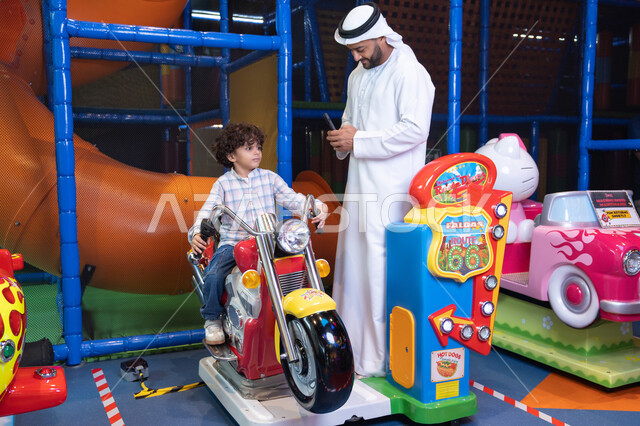 التفاعل بين أفراد العائلة، الأنشطة الرياضية الممتعة، أجواء المرح والحماس، طفل عربي خليجي يركب لعبة الدراجه المتحركه بايماءات السعاده و السرور ، رجل عربي خليجي اماراتي يرتدي الكندورة و الغتره يستمتع بقضاء وقت ممتع مع ابنه فى مدينه الملاهي