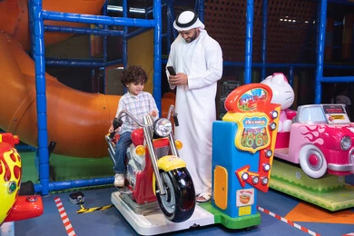 الأنشطة الرياضية الممتعة، التفاعل بين أفراد العائلة ، أجواء المرح والحماس، طفل عربي خليجي يركب لعبة الدراجه المتحركه بايماءات السعاده و السرور ، رجل عربي خليجي اماراتي يرتدي الكندورة و الغتره يستمتع بقضاء وقت ممتع مع ابنه فى مدينه الملاهي