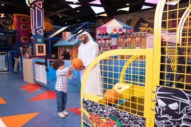 Entertainment center, an Emirati Gulf Arab man wearing a kandura and ghutra enjoying a fun time with his child in an amusement park, a father sharing moments of fun and excitement with his son, a cheerful interactive atmosphere, expressions of pleasure and happiness, a Gulf child wearing casual clothes playing basketball, recreational sports activities for children