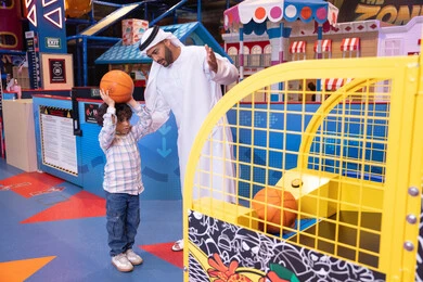 A cheerful interactive atmosphere, expressions of pleasure and happiness, a Gulf child wearing casual clothes playing basketball, recreational sports activities for children, a father sharing moments of fun and excitement with his son, an Emirati Gulf Arab man wearing a kandura and ghutra enjoying a fun time with his child in an amusement park