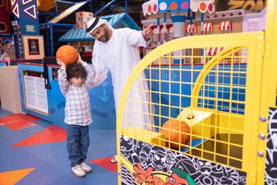 تعابير السرور والسعادة، طفل خليجي يرتدي الزي الكاجوال يقوم  بلعب كره السله، انشطة رياضية ترفيهية للاطفال، مشاركة الأب ابنه لحظات المرح والحماس ، أجواء تفاعلية مبهجة ، رجل عربي خليجي اماراتي يرتدي الكندورة و الغتره يستمتع بقضاء وقت ممتع مع طفله فى مدينه الملاهي