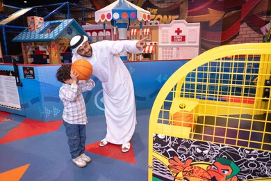 طفل خليجي يرتدي الزي الكاجوال يقوم  بلعب كره السله، انشطة رياضية ترفيهية للاطفال،  تعابير السرور والسعادة، مشاركة الأب ابنه لحظات المرح والحماس ، أجواء تفاعلية مبهجة ، رجل عربي خليجي اماراتي يرتدي الكندورة و الغتره يستمتع بقضاء وقت ممتع مع طفله فى مدينه الملاهي