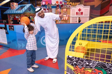 انشطة رياضية ترفيهية للاطفال، طفل خليجي يرتدي الزي الكاجوال يقوم  بلعب كره السله،  تعابير السرور والسعادة، مشاركة الأب ابنه لحظات المرح والحماس ، أجواء تفاعلية مبهجة ، رجل عربي خليجي اماراتي يرتدي الكندورة و الغتره يستمتع بقضاء وقت ممتع مع طفله فى مدينه الملاهي