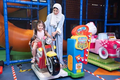 طفلة خليجيه ترتدي الزي الكاجوال تركب لعبة الدراجة بايماءات السعادة و السرور، مشاركة الاطفال بالانشطة الترفيهية، ام عربيه خليجيه اماراتية ترتدي العباءه الملونه تقوم بقضاء وقت ممتع مع طفلتها فى مدينة الالعاب، العاب الكترونية ، التسلية و الترفيه