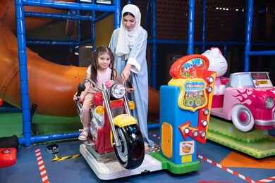 Children participating in recreational activities, an Emirati Gulf Arab mother wearing a colorful abaya is spending a fun time with her daughter in the amusement park, electronic games, entertainment and recreation, a Gulf girl wearing casual clothes is riding a bicycle with gestures of happiness and pleasure
