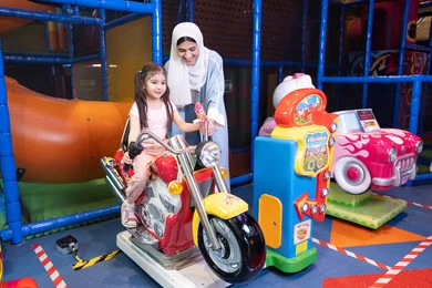 أجواء تفاعلية مبهجة، ام عربيه خليجيه اماراتية ترتدي العباءه الملونه تقوم بقضاء وقت ممتع مع طفلتها فى مدينة الالعاب، العاب الكترونية ، التسلية و الترفيه ، طفلة خليجيه ترتدي الزي الكاجوال تركب لعبة الدراجة بايماءات السعادة و السرور