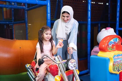 الشعور بالمتعة والفرح، ام عربيه خليجيه اماراتية ترتدي العباءه الملونه تقوم بقضاء وقت ممتع مع طفلتها فى مدينة الالعاب، العاب الكترونية ، التسلية و الترفيه ، طفلة خليجيه ترتدي الزي الكاجوال تركب لعبة الدراجة بايماءات السعادة و السرور