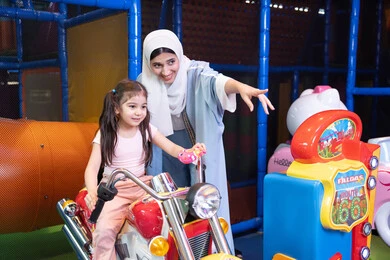 أجواء مليئة بالمرح والحماس، ام عربيه خليجيه اماراتية ترتدي العباءه الملونه تقوم بقضاء وقت ممتع مع طفلتها فى مدينة الالعاب، العاب الكترونية ، التسلية و الترفيه ، طفلة خليجيه ترتدي الزي الكاجوال تركب لعبة الدراجة بايماءات السعادة و السرور