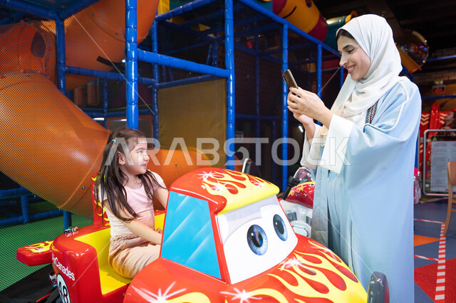 Children participating in recreational activities, documenting happy moments, improving the quality of life using modern technology, an Emirati Gulf Arab woman wearing a colorful abaya spending time with her daughter, recreational summer activities, playing recreational games, spending fun family time, a happy summer vacation, a Saudi mother taking a souvenir photo with her mobile phone