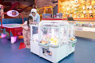 Summer vacation entertainment, enjoying playing with children, spending the weekend in places of fun and entertainment, an Emirati Gulf Arab mother wearing a colorful abaya spending a fun time with her daughter in an amusement park, a Gulf girl playing on the claw machine with gestures of happiness and pleasure, electronic games