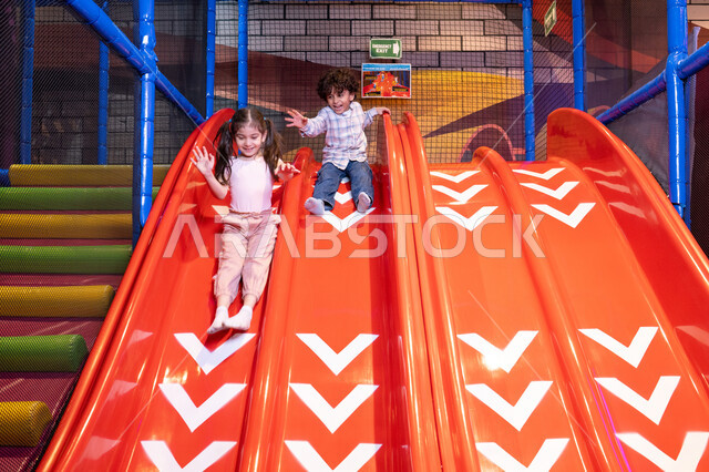 Children participating in recreational activities, sliding on the slide, gestures of joy and happiness, two Emirati Gulf Arab children wearing casual clothes enjoying playing on the slide, exploiting free time in entertainment and play