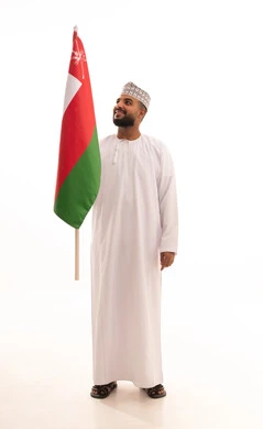 Pride and honor in the symbols and flag of Oman, participation in national holidays and events, celebrating the anniversary of Oman's National Day, a portrait of a young Arab Gulf Omani man wearing a dishdasha and kumma holding the Omani flag in his hands, the concept of love for the homeland and belonging to it, full-body, white background.