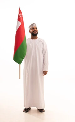 Participation in national holidays and events, celebrating the anniversary of Oman's National Day, pride and honor in Omani symbols and the flag, a portrait of a young Arab Gulf Omani man wearing a dishdasha and kumma holding the Omani flag in his hands, the concept of love for the homeland and belonging to it, full-body image, white background.