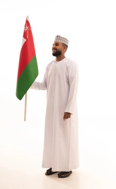 Celebrating the anniversary of Oman's National Day, participating in national holidays and events, pride and honor in Omani symbols and the flag, a portrait of a young Arab Gulf Omani man wearing a dishdasha and kumma holding the Omani flag in his hands, the concept of love for the homeland and belonging to it, full-body, white background.
