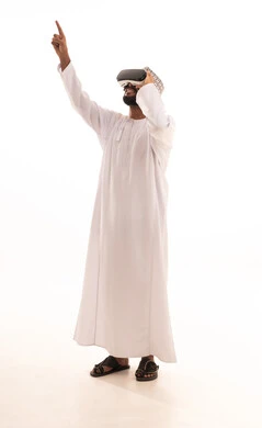 التفاعل والاندماج مع الواقع الافتراضي، استخدام الأجهزة التقنية الحديثة، الاشارة بإصبع السبابة إلى شيء ما، رؤية المشاهد الخيالية،بورتريه لشاب عربي خليجي عماني يرتدي الدشداشة والكمه يضع نظارات الفي آر، قضاء أوقات الفراغ بشكل ممتع، الجسم بالطول الكامل، خلفية بيضاء