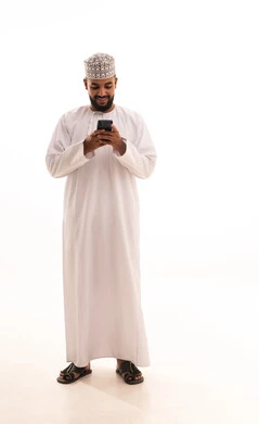 Browsing mobile applications, chatting and talking with friends online, a portrait of a young Arab Gulf Omani man wearing a dishdasha and kumma, following social media news using a mobile phone, gestures of happiness and joy, full-body image, white background.