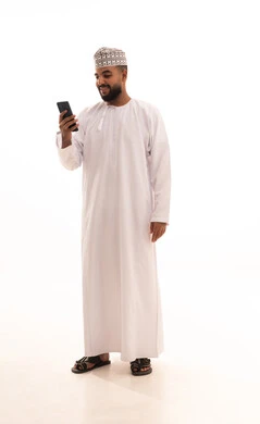 الدردشة والتحدث مع الاصدقاء عبر الانترنت،بورتريه لشاب عربي خليجي عماني يرتدي الدشداشة والكمه يقوم بمتابعة اخبار وسائل التواصل الاجتماعي باستخدام الجوال، تصفح تطبيقات الهاتف المحمول، ايماءات السعادة والسرور، صورة الجسم بالطول الكامل، خلفية بيضاء