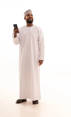 Communicating with family and friends via mobile, using advanced technological devices, a portrait of a young Arab Gulf Omani man wearing a dishdasha and kumma holding a mobile phone in his hand and looking at something, a full-body image, white background.