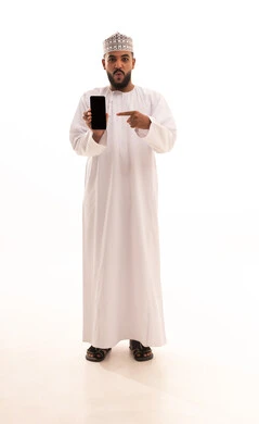 النظر إلى الكاميرا بايماءات المفاجئة والتعجب، عرض شاشة سوداء فارغة على الهاتف المحمول، استخدام الوسائل التقنية الحديثة، بورتريه لشاب عربي خليجي عماني يرتدي الدشداشة والكمه يحمل الجوال ويشير اليه بإصبع السبابة، صورة لكامل الجسم، خلفية بيضاء