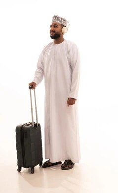 الاستعداد لقضاء رحلة صيفية ممتعة, الذهاب في جولات ترفيهية في إجازة الصيف, تعابير الفرح والسعادة، بورتريه لشاب عربي خليجي عماني مبتسم يرتدي الدشداشة و الكمة و الهيدفون يمسك حقيبة السفر ينظربإيماءات السرور, صورة الجسم بالطول الكامل، خلفية بيضاء