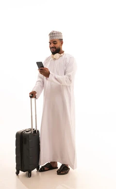 تصفح تطبيقات الهاتف المحمول، التحدث والدردشة مع الاصدقاء عبر الانترنت، الاستعداد لقضاء رحلة صيفية ممتعة، بورتريه لشاب عربي خليجي عماني يرتدي الدشداشه والكمه يمسك بيديه حقيبة السفر يقوم بمتابعة اخبار وسائل التواصل الاجتماعي ، الجسم بالطول الكامل، خلفية بيضاء