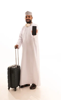 حجز اونلاين عبر تطبيقات وبرامج السياحة, بورتريه لشاب عربي خليجي عماني مبتسم يرتدي الدشداشة والكمة  يمسك بيدية حقيبة السفر و الهاتف المحمول, الاستعداد لقضاء رحلة صيفية ممتعة, عرض شاشة سوداء فارغة، صورة الجسم بالطول الكامل، خلفية بيضاء