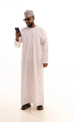 التقاط صور سيلفي تذكارية عبر الجوال، اجراء مكالمة فيديو عبر تطبيقات ومواقع السوشال ميديا، استخدام الأجهزة والتقنيات الحديثة، بورتريه لشاب عربي خليجي عماني يرتدي الدشداشة والكمه والنظارة الشمسية يقوم بحمل الهاتف المحمول، ايماءات الدهشة والسرور، الجسم بالطول الكامل، خلفية بيضاء