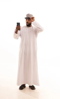 اجراء مكالمة فيديو عبر تطبيقات ومواقع السوشال ميديا، التقاط صور سيلفي تذكارية عبر الجوال، استخدام الأجهزة والتقنيات الحديثة، بورتريه لشاب عربي خليجي عماني يرتدي الدشداشة والكمه والنظارة الشمسية يقوم بحمل الهاتف المحمول، ايماءات الدهشة والسرور، الجسم بالطول الكامل، خلفية بيضاء