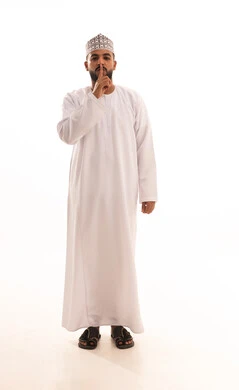 الأمر بعدم الازعاج والسكوت والصمت، بورتريه لشاب عربي خليجي عماني يرتدي الدشداشة والكمه يضع اصبع السبابة على فمه ينظر الى الكاميرا بإيماءات التوقف عن الكلام وطلب خفض الصوت، تعابير الجدية والحزم والغضب، صورة الجسم بالطول الكامل، خلفية بيضاء