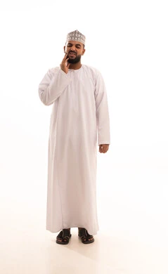 The concept of tooth decay, a portrait of a young Arab Gulf Omani man wearing a dishdasha and kumma, placing his hand on his face with gestures of pain and toothache, swelling and inflammation in the mouth and gums, the necessity to go to the doctor and receive treatment, a full-body image, white background.