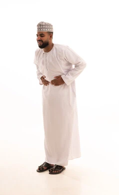 الإحساس بالمغص و التعب والإرهاق, الحاجة للدواء ووضع خطة علاجية, عسر الهضم والشعور بحرقة في المعدة، بورتريه لشاب عربي خليجي عماني يرتدي الدشداشة والكمه يضع يديه على بطنه بإيماءات الألم، صورة الجسم بالطول الكامل، خلفية بيضاء