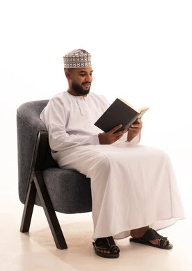 التزود بالمعلومات من الكتب والروايات، مطالعة القصص الممتعة، مفهوم التعلم الذاتي، تعابير التمعن والتركيز، قضاء وقت الفراغ بالقراءة والتعلم والثقافة،بورتريه لرجل عربي خليجي عماني يرتدي الدشداشة والكمة يجلس على كرسي يقوم بمطالعة كتاب، خلفية بيضاء