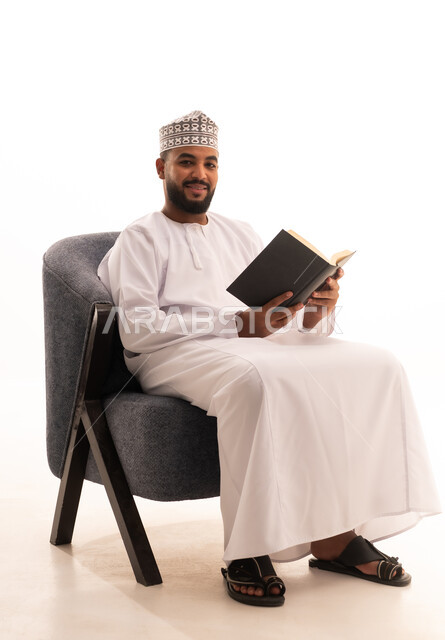 ايماءات السعادة و السرور، مطالعة القصص الممتعة، مفهوم التعلم الذاتي، قضاء وقت الفراغ بالقراءة والتعلم والثقافة،بورتريه لرجل عربي خليجي عماني يرتدي الدشداشة والكمة يجلس على كرسي يقوم بمطالعة كتاب، التزود بالمعلومات من الكتب والروايات، خلفية بيضاء