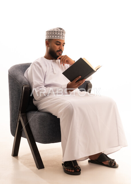 مطالعة القصص الممتعة، مفهوم التعلم الذاتي، تعابير التمعن والتركيز، قضاء وقت الفراغ بالقراءة والتعلم والثقافة،بورتريه لرجل عربي خليجي عماني يرتدي الدشداشة والكمة يجلس على كرسي يقوم بمطالعة كتاب، التزود بالمعلومات من الكتب والروايات، خلفية بيضاء