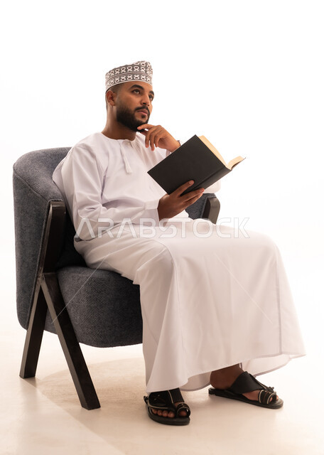 Gestures of thinking about something, reading interesting stories, the concept of self-learning, spending free time reading, learning, and culture, a portrait of an Arab Gulf Omani man wearing a dishdasha and kumma sitting on a chair reading a book, acquiring information from books and novels, white background.