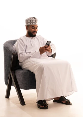 Browsing social media, using a modern technical device, communicating with family and friends, making a video call via mobile, a portrait of an Arab Gulf Omani man wearing a dishdasha and kumma sitting on a chair and working on a mobile phone, white background.