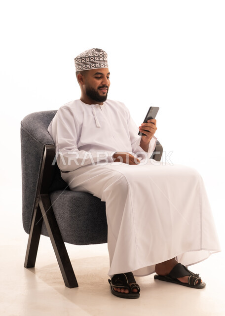Using a modern technical device, communicating with family and friends, making a video call via mobile, browsing social media, a portrait of an Arab Gulf Omani man wearing a dishdasha and kumma sitting on a chair and working on a mobile phone, white background.