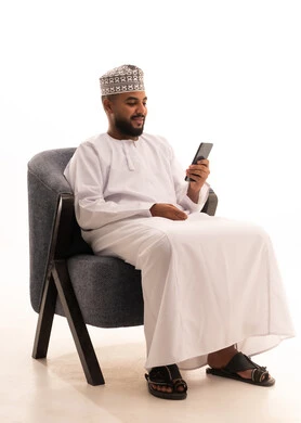 Using a modern technical device, communicating with family and friends, making a video call via mobile, browsing social media, a portrait of an Arab Gulf Omani man wearing a dishdasha and kumma sitting on a chair and working on a mobile phone, white background.