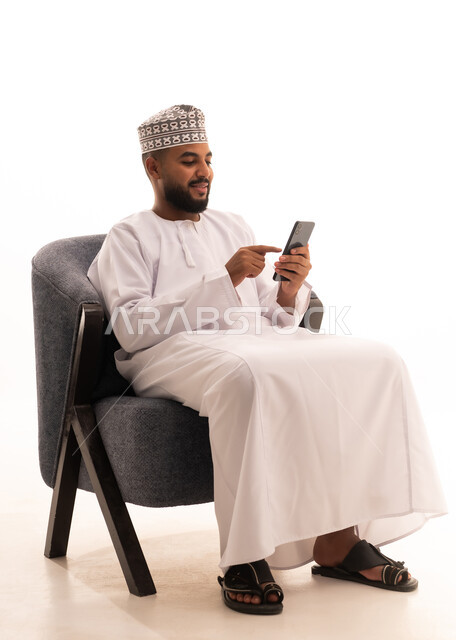 التواصل مع الأهل والاصدقاء، إجراء مكالمة فيديو عبر الجوال، تصفح مواقع التواصل الاجتماعي، استخدام جهاز تقني حديث،بورتريه لرجل عربي خليجي عماني يرتدي الدشداشة والكمة يجلس على كرسي ويعمل على الهاتف المحمول، خلفية بيضاء