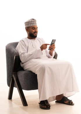 Communicating with family and friends, making a video call via mobile, browsing social media, using modern technology, a portrait of an Arab Gulf Omani man wearing a dishdasha and kumma sitting on a chair and working on a mobile phone, white background.